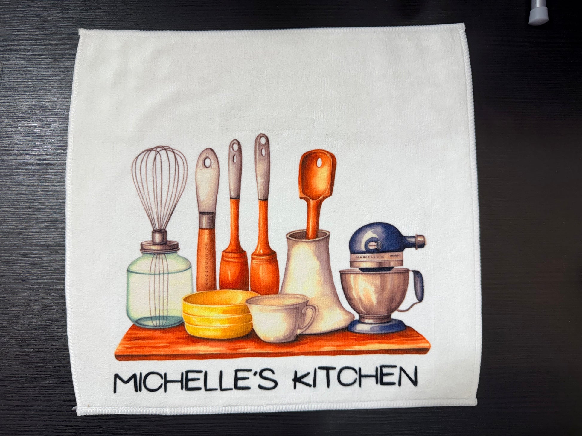 Kitchen towel with a design of kitchen utensils and the text 'Michelle's Kitchen' on a dark surface.